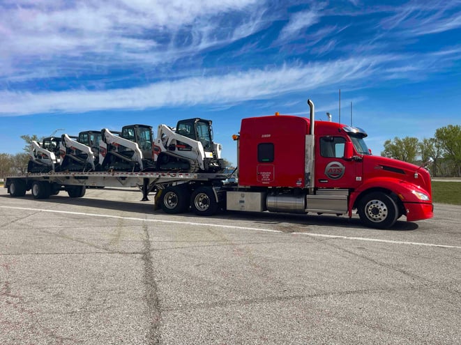 A red ATS truck hauls a flatbed trailer loaded with several pieces of construction equipment.
