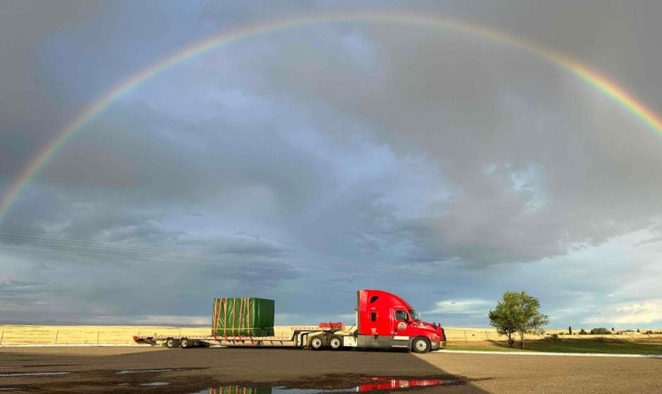 Semi truck and loaded flatbed trailer parked beneath rainbow in cloudy sky