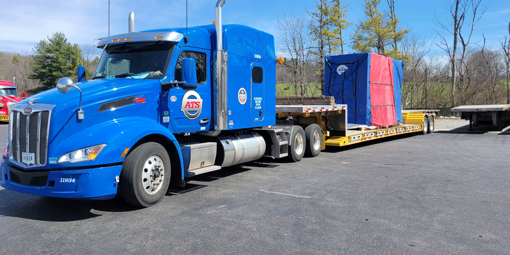 Blue ATS truck hauling a step-deck trailer with a tarped load.