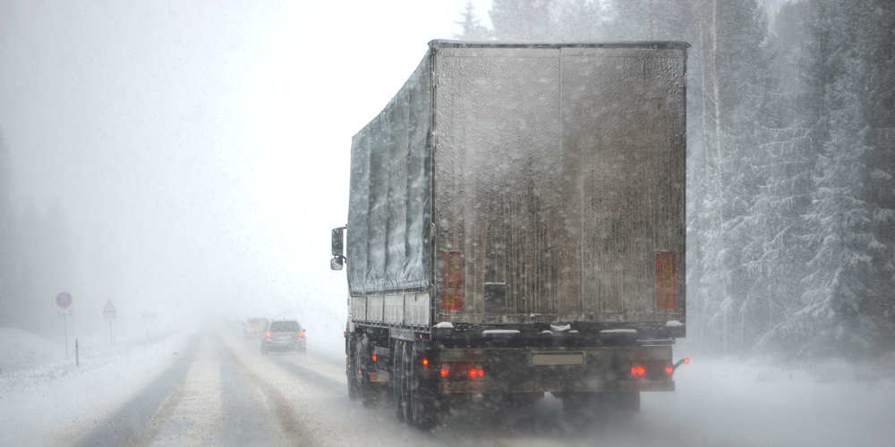 Truck driving on a two-lane, snow-covered highway surrounded by snowy pine trees.