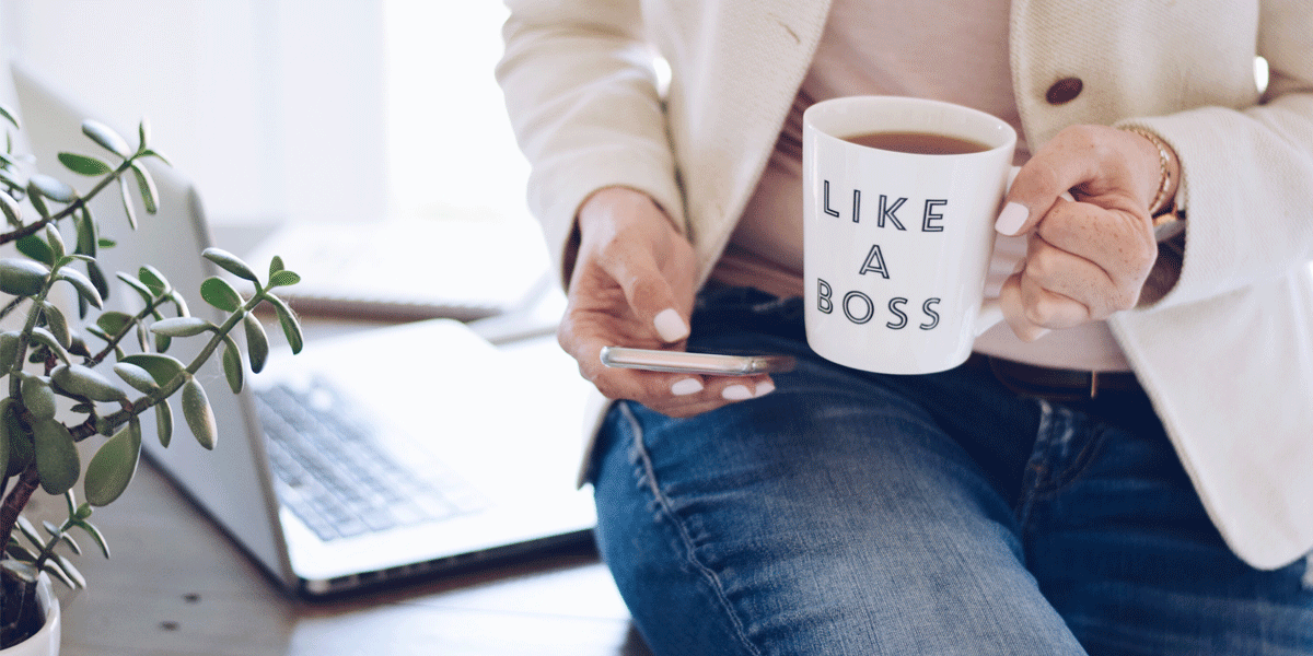 Woman sitting on a desk with a plant and laptop. She looks at her phone as she holds a mug that says "Like a Boss."