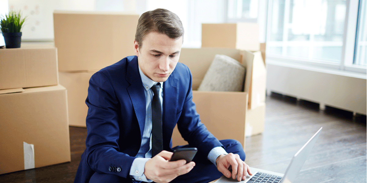 A young man in blue suit surrounded by boxes. He's on his phone and his laptop sits opened nearby.