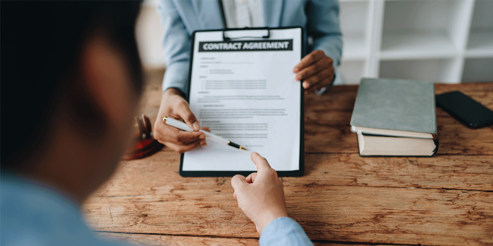 A person behind a desk pointing at where to sign on a sheet of paper.