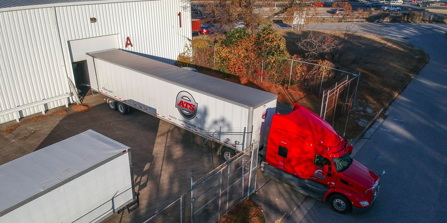 Red truck with dry van trailer backing into manufacturing facility